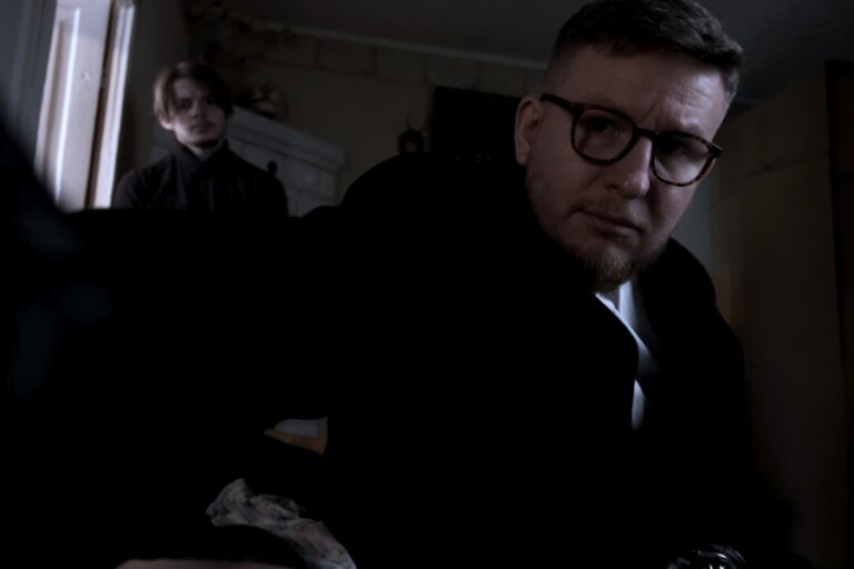 Kadr z filmu "Cena Duszy". Mężczyzna na pierwszym planie trzyma kamerę w dłoni i patrzy w obiektyw. W tle drugi mężczyzna również patrzy w stronę kamery.