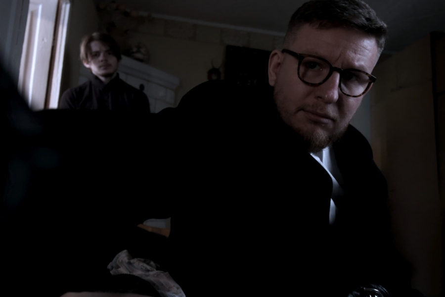 Kadr z filmu "Cena Duszy". Mężczyzna na pierwszym planie trzyma kamerę w dłoni i patrzy w obiektyw. W tle drugi mężczyzna również patrzy w stronę kamery.