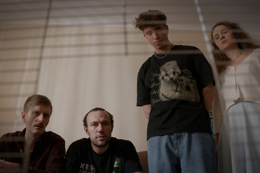Kadr z filmu "DAMMIT". Cztery osoby patrzą w obiektyw kamery umieszczonej w klatce dla małego zwierzęcia.
