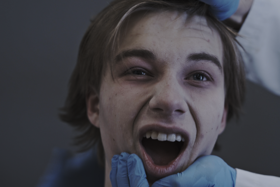 Kadr z filmu "Pchły w słoiku". Osoba z szeroko otwartymi ustami, trzymana za szczękę przez ręce w niebieskich rękawiczkach.