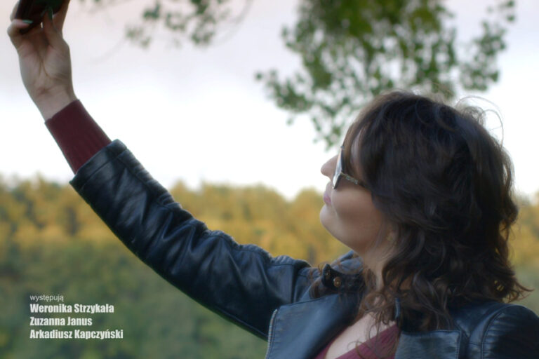 Fragment plakatu do filmu "Sobota". Kobieta robi sobie selfie na zewnątrz, w tle drzewa.
