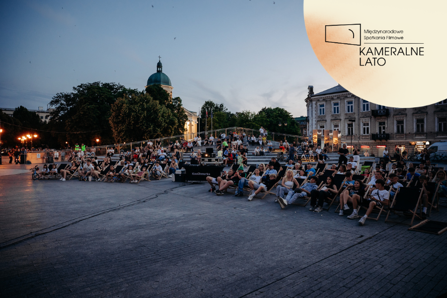 Publiczność siedzi na leżakach podczas plenerowego wydarzenia filmowego na miejskim placu o zmierzchu. W prawym górnym rogu widoczne logo Kameralnego Lata.