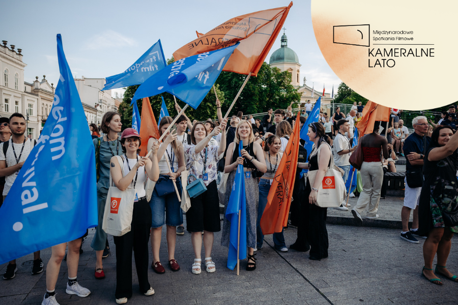 Uczestnicy parady festiwalowej stoją na placu z niebieskimi i pomarańczowymi flagami, uśmiechając się do zdjęcia. W prawym górnym rogu widoczne logo Kameralnego Lata.