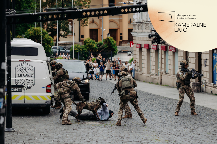 Inscenizowana scena z udziałem funkcjonariuszy w umundurowaniu taktycznym i psa służbowego podczas kręcenia sceny specjalnej w Radomiu. Na brukowanej ulicy obezwładniana jest osoba, a w tle przyglądają się widzowie.